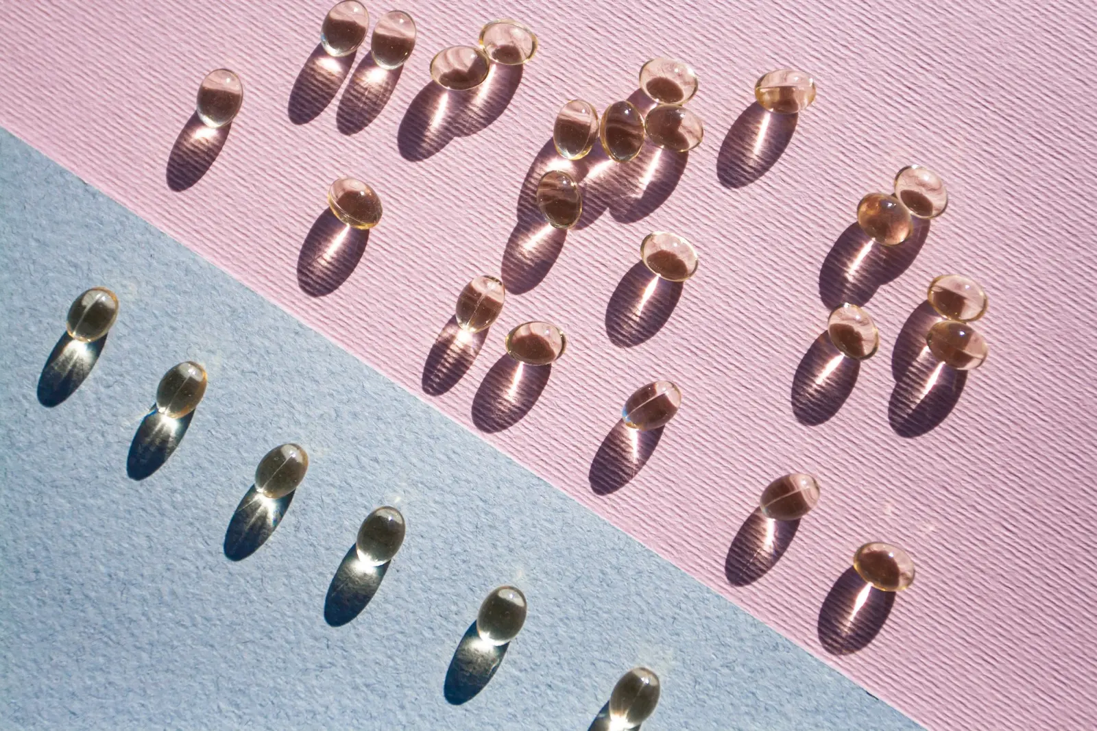 Amber softgel capsules arranged on pink and blue paper in raking sunlight